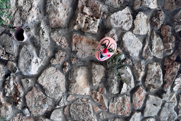 A discarded, pink clown mask with a wide smile and red nose is positioned on a rugged, irregularly shaped stone wall.