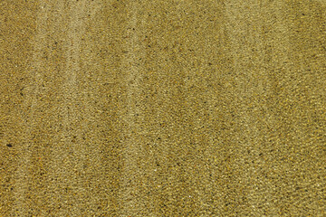 Clear speckled water flows over sandy riverbed covered with evenly spaced medium sized rock. Midday sunlight adds shimmering web-like surface to river ripples thanks to lacking bottom vegetation.