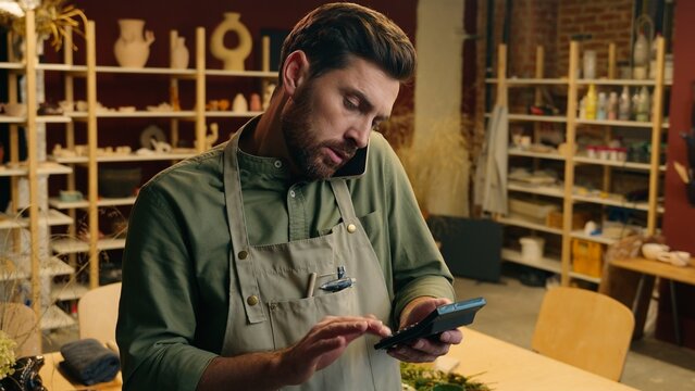 Busy Caucasian man male guy small business owner businessman potter craftsman using calculator counting finance calculate price money talking mobile smartphone phone call client order inside workshop