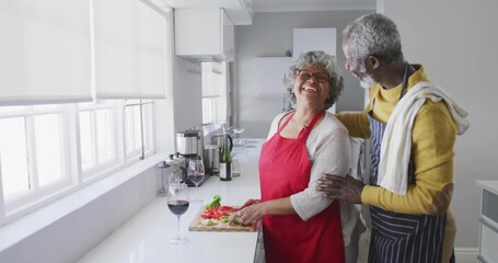 Senior couple in aprons slicing tomatoes together on cutting board in kitchen, with wine glasses