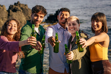 Diverse group of friends looking at camera toasting with beer on beach at sunset. Concept of gen z celebration, multi-ethnic friendship, summer vacation party, and making a toast to the viewer (POV).