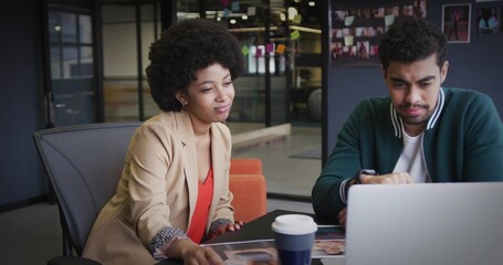 Leaning coworkers wearing blazer and bomber jacket examining photos in modern office, with laptop
