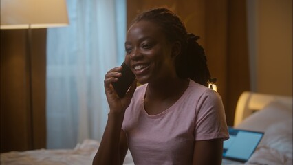 Laughing cheerful African American ethnic woman female lady girl holding mobile phone chatting cellphone conversation talk smartphone call gossip friends sitting bed indoor home bedroom late evening