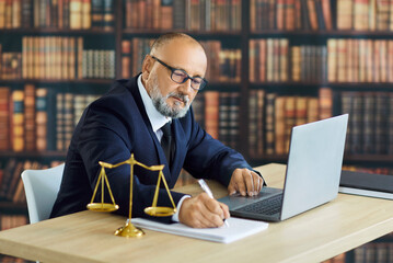 Lawyer attorney uses laptop beside scales. In a law library office, legal advisor takes notes and...