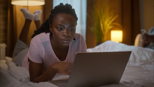 Family weekend relaxed calm African American woman girlfriend browsing internet typing laptop computer lying on bed man boyfriend chatting mobile phone texting gadget addiction at home bedroom evening