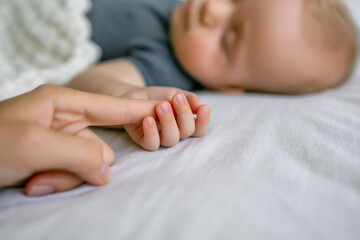 Close up loving mommy holds hand of her newborn while baby sleeping lying on bed during healthy day...