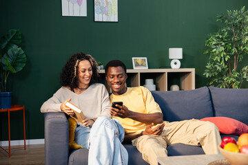Happy black couple relaxing on sofa using smartphone
