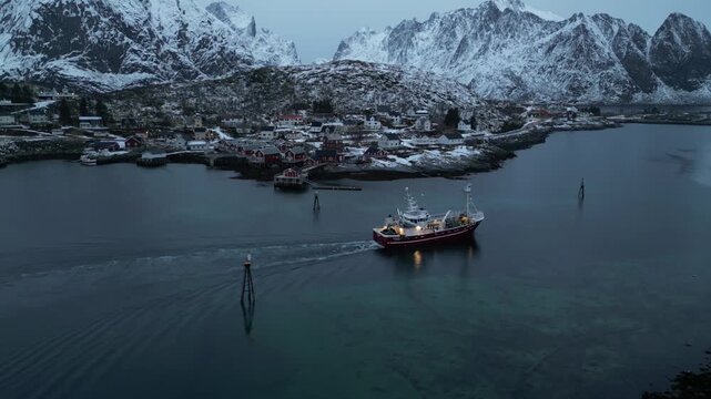 Aerial view of a fishing boat sailing through calm waters near Reine, where red houses nestle against snow-covered mountains, Lofoten Islands, Norway.