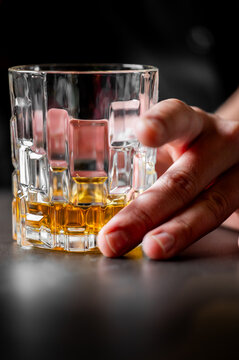 A hand reaches for a crystal whiskey glass filled with amber-colored liquid and ice on a dark table. The glass features a geometric, textured pattern inspired by natural imperfections.