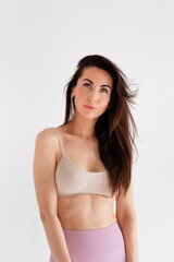 Fototapeta premium Sports woman standing in activewear with abs muscles, looking at camera isolated on white background. Caucasian 30s fitness girl wears beige top and pink leggings. Slim fit brunette with blue eyes.