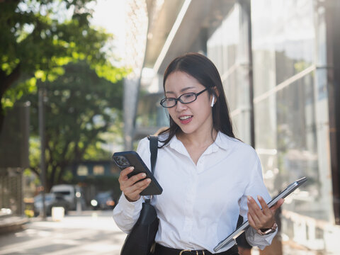Busy young Asian businesswoman multitasking, talking on a wireless earbud while checking her smartphone and carrying a laptop, walking quickly through the city