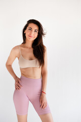 Fototapeta premium Sports woman standing in activewear with abs muscles, looking at camera isolated on white background. Caucasian 30s fitness girl wears beige top, pink leggings. Slim fit smiling brunette, blue eyes.