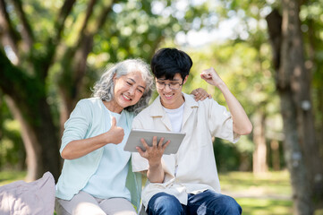 Fototapeta premium Thumb up old woman grandmother looking at tablet and young man grandson raising a fist sits in park.