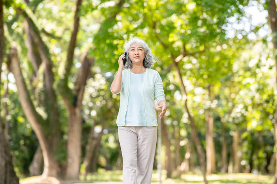 Old asian woman grandma holding and talking on smartphone while strolling or walking in the park. - Powered by Adobe