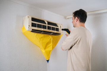 Air conditioning technician cleaning air conditioner unit using steam cleaner. While yellow protective bag collects dirty water. Ensuring thorough and professional cleaning process.