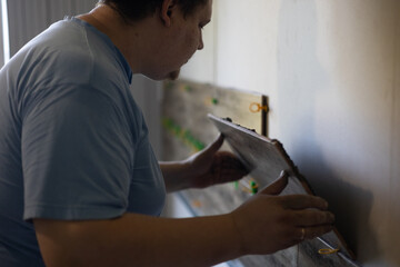 Obraz premium A man lays ceramic tiles on an interior wall while renovating or improving a home. The process of laying tiles symbolizes independent projects and skilled labor. Laying tiles on a wall in a house.