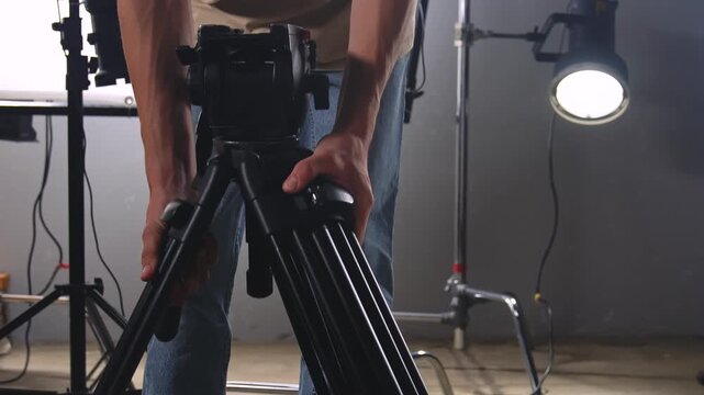 Close up of a man setting up a tripod in a photo and video studio with professional lighting. Preparing for a shoot or photo session in a creative workspace.