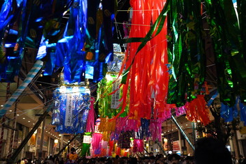 夜の商店街の頭上に赤青緑など極彩色の七夕飾りが密集して吊るされお祭りの熱気と賑やかさを感じさせます