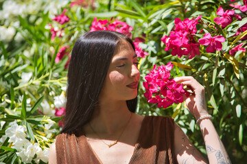 Fototapeta premium Portrait of young woman, brunette, tall, thin and very tattooed, wearing a brown dress, with her eyes closed, smelling the perfume of pink oleanders. Concept of youth, rebelliousness, millennial.