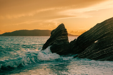 Dramatic coastal scene with ocean waves crashing against sharp rock formations at sunset, under a warm golden sky