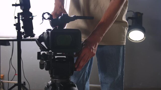 A man in casual clothes sets up and adjusts a professional video camera on a tripod in a film studio with lighting equipment. Off-screen video production setup process.
