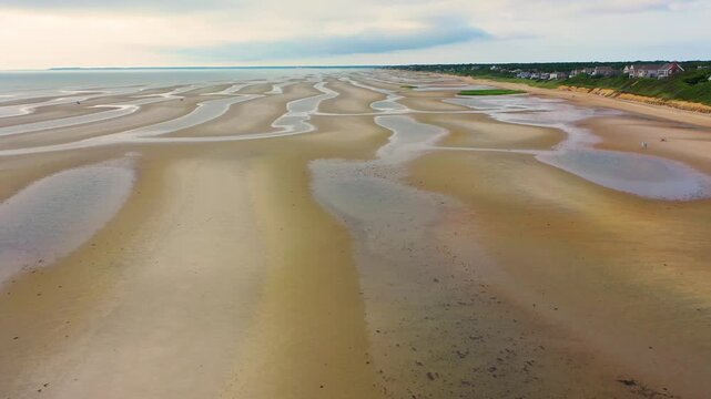 Aerial view of a beach at low tide reveals flowing sandbars, shallow tide pools glistening in the sun, and natural dune-like patterns stretching across the shoreline beneath open skies.