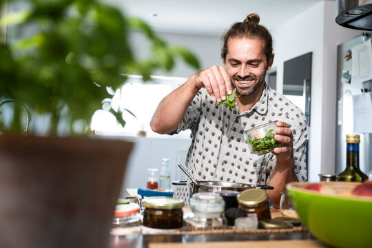 Happy man adding basil leaves and cooking food in kitchen at home
