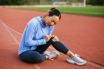 Female athlete holding painful knee sitting on outdoor stadium running track