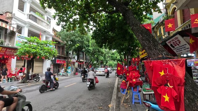 A lively street scene in Hanoi Old Quarter, Vietnam with motorbikes passing by shops decorated with Vietnamese flags and trees lining the road. Panning footage.