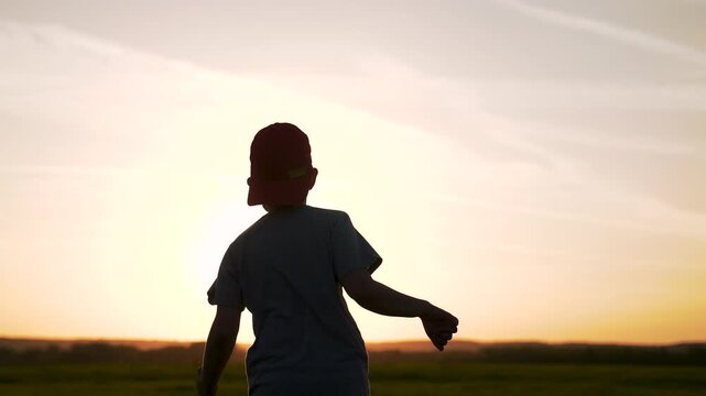 Child running toward sunset silhouette as sun dips over field with cap backward backlight creating dark figure against glowing horizon outdoor showing motion and playful energy of running child