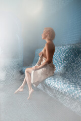 Serene woman in white towel sitting in profile on blue mosaic bench surrounded by thick steam and mist in tranquil hammam spa, soft diffused lighting creating wellness atmosphere and relaxation