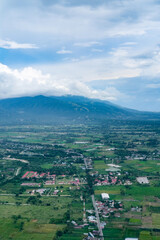Fototapeta premium Majestic aerial view of a mountain valley surrounded by lush hills and clouds, overlooking a sprawling town below with green fields, scattered houses, and winding roads blending into the landscape.