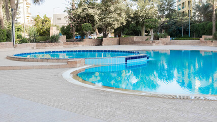 A serene swimming pool with clear blue water, surrounded by palm trees and landscaped gardens. The pool features a curved design and tiled edges.