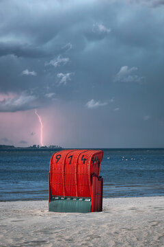 A red beach chair on a sandy beach of the Baltic Sea in the Kiel Fjord during a thunderstorm with a flash of lightning.