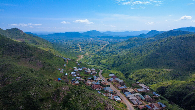 Aerial view of houses nestled between rolling green hills under a vast blue sky, a tranquil contrast of nature and habitation, Jos, Plateau, Nigeria.