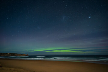 The green southern lights of the Aurora Australis over the dark, blue ocean as waves break on the sandy beach. This spectacular display is seen from the coast of Phillip Island, Australia.