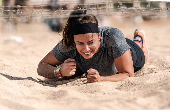 Strong female athlete smiling and enjoying crawling under barbed wire on an obstacle course in the sand during an intense obstacle course race at Santa Susanna beach, Barcelona.