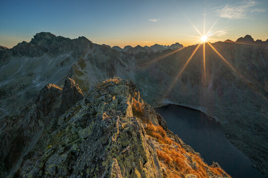 Fototapeta Aerial view of jagged peaks meet the sky as the sun bursts forth, casting golden rays over the serene lake below, Vysoke Tatry, Presovsky kraj, Slovakia.