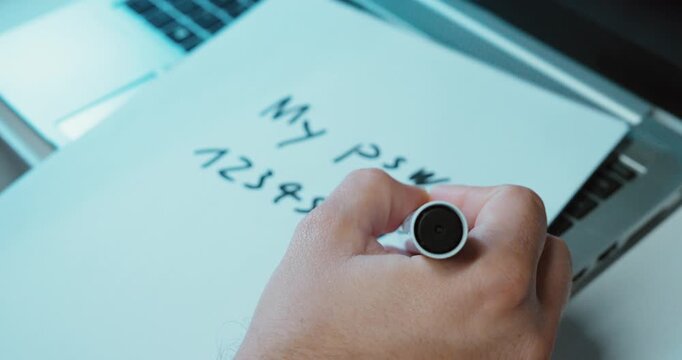 A person writes down their password on paper next to a laptop. Concept: cybersecurity awareness, phishing, password protection, 2FA, company training, data breach prevention.