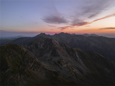 Aerial view of jagged, rocky mountain peaks pierce the twilight sky, kissed with hues of orange and purple, a vast wilderness unfolds below., Bucharest, Romania.
