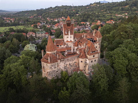 Aerial view of Bran Castle's imposing silhouette rises from a sea of emerald trees, its terracotta rooftops contrasting with the stone walls, Bran, Romania.