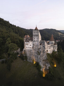 Aerial view of Bran Castle perched atop a rocky precipice, its aged stone walls contrasting with the verdant forest, creating a timeless scene, Bran, Romania.