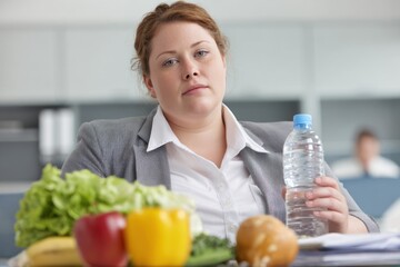 A woman in business attire sits at a table with fruits and vegetables, holding a water bottle, appearing contemplative or stressed in a modern office setting.