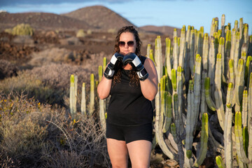 Young woman wearing black sleeveless athletic top, black shorts and boxing gloves in black and white holding her fists raised in a defensive stance, centered posture showing determination and strength