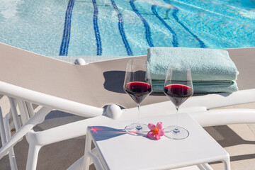 Two glasses of red wine placed on a white side table beside a comfortable sun lounger near a crystal clear swimming pool in a holiday resort setting, display of luxury, leisure and holiday relaxation
