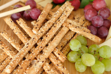 Wooden serving board featuring a pile of crispy sesame breadsticks, fresh red and green grapes, and a variety of sliced cheeses, creating a rustic tapas platter perfect for a party or social gathering