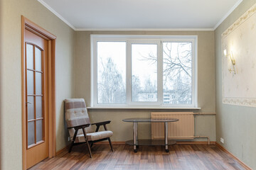 an old living room without renovation. There's old wallpaper on the walls. The room needs renovation. near the large window there is a battered armchair and a glass coffee table