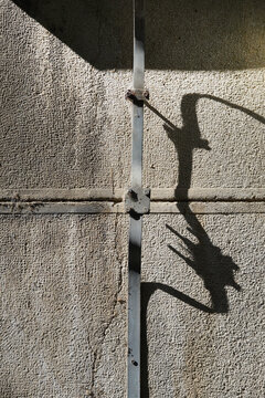 A weathered, textured concrete wall features a vertical lightning rod strip casting sharp, abstract shadows in harsh sunlight