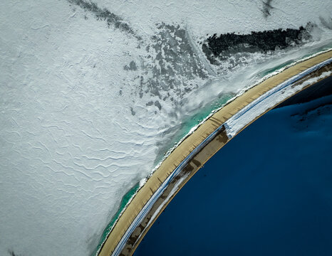 Aerial view of a road cutting through the stark white expanse of a frozen lake contrasted by the deep blues of open water, Lucomagno, Ticino, Switzerland.