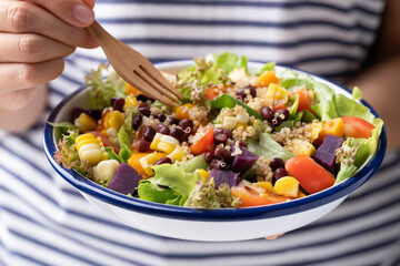 Healthy vegetable salad with lettuce, quinoa seed, azuki bean, corn, tomato, pumpkin and purple sweet potato in a bowl holding by woman hand and eating. Vegan and plant-based food
